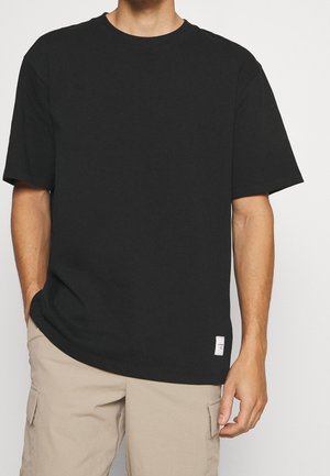 Hombre vestido con una camiseta negra de mangas cortas sin estampado, que tiene una pequeña etiqueta blanca en el dobladillo inferior, y pantalones cortos cargo beige, con las manos relajadas a los lados.