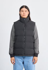 Gilet noir matelassé avec col montant, fermeture éclair et design matelassé ; porté par-dessus une chemise à manches longues gris à motifs. Ajustement modifiable.