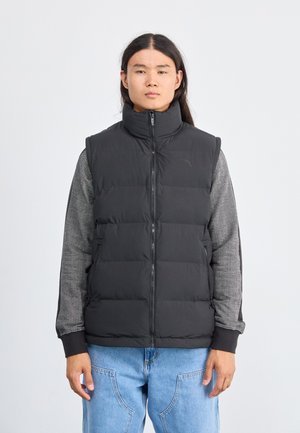 Gilet noir matelassé avec col montant, fermeture éclair et design matelassé ; porté par-dessus une chemise à manches longues gris à motifs. Ajustement modifiable.