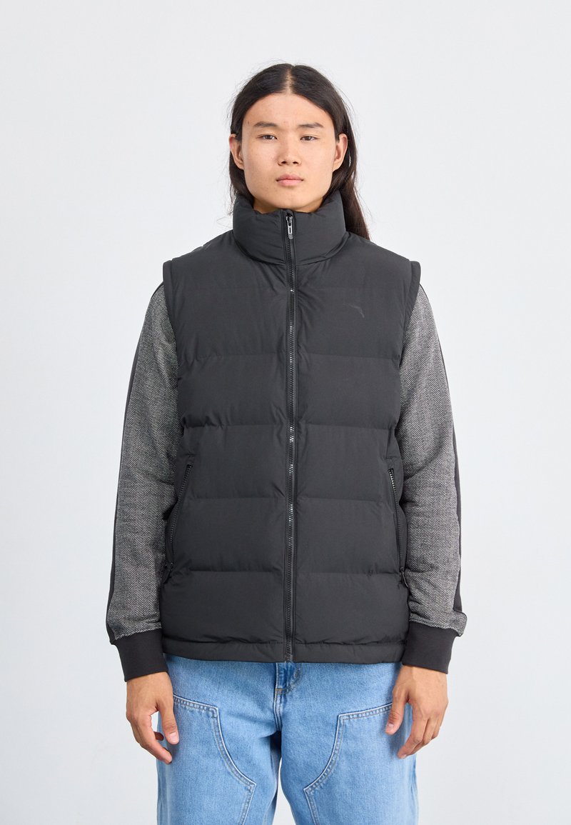 Gilet noir matelassé avec col montant, fermeture éclair et design matelassé ; porté par-dessus une chemise à manches longues gris à motifs. Ajustement modifiable.