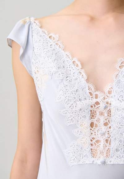 Donna che indossa un vestito azzurro pallido con dettagli intricati di pizzo floreale bianco e piccoli bottoni sul petto e sulle maniche a cappuccio.