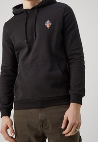 Felpa nera con un grafico a forma di diamante sul petto, polsini a coste, tasca frontale e cappuccio con cordoncino. Tessuto morbido.