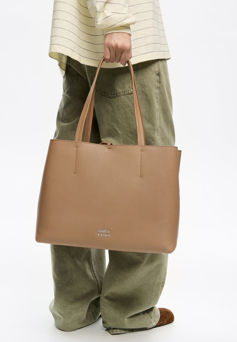 Bolso tote de cuero marrón claro con dos asas largas, que presenta una superficie texturizada y un pequeño logo en la parte inferior. Se lleva con pantalones olivo relajados.