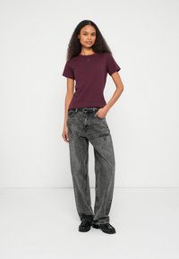 Μπορντό κοντομάνικη μπλούζα με στρογγυλή λαιμόκοψη και μικρό λογότυπο στο στήθος, συνδυασμένη με ψηλόμεσα, φαρδιά γκρι τζιν και μαύρα παπούτσια.