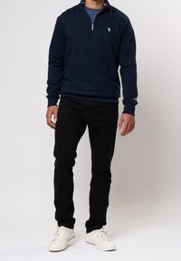 Mörkblå sweatshirt med dragkedja vid halsen och långa ärmar, med en liten logotyp på bröstet. Kombinerad med svarta slim-fit-byxor och vita sneakers.