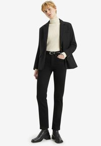 Blazer negro ajustado sobre un suéter de cuello alto de canalé en color crema, combinado con jeans ajustados negros y botas de tobillo negras. Un atuendo simple y monocromático.