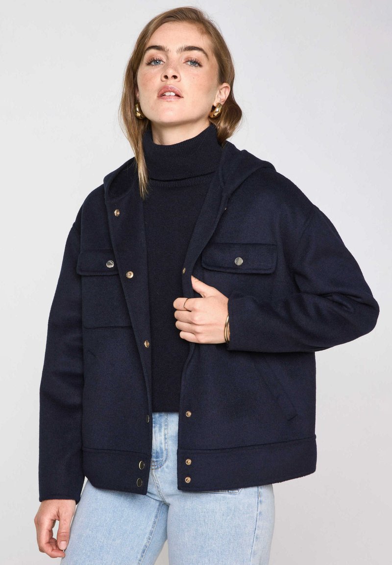 Giacca blu navy con cappuccio, realizzata in tessuto morbido. Presenta due tasche sul petto, bottoni dorati e una vestibilità rilassata. Indossata sopra un dolcevita.