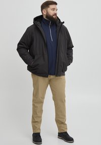 Jachetă neagră impermeabilă cu glugă, cu fermoar pe față și două buzunare frontale, purtată peste un pulover bleu marin. Pantaloni bej și pantofi negri completează ținuta.