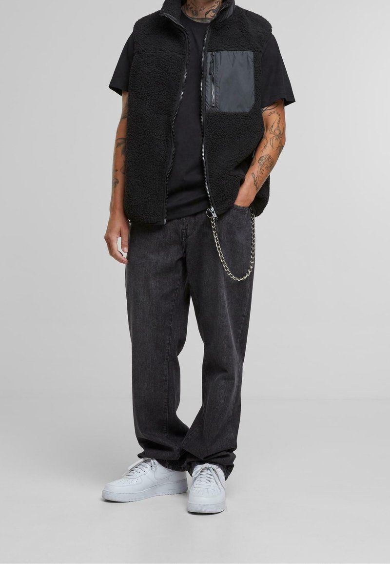 Czarna kamizelka polarowa z kieszenią na zamek, zestawiona z czarną koszulką i ciemnoszarymi spodniami z denimu. Obuwie to białe trampki z perforacjami.