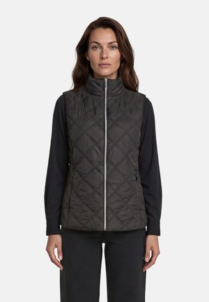 Femme portant un gilet matelassé noir avec un col montant et une fermeture éclair frontale sur une chemise noire à manches longues, debout devant un fond blanc uni.