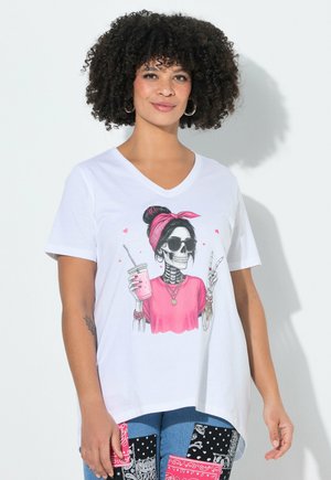 Vrouw met krullend haar die een wit V-hals T-shirt draagt met een skelet in zonnebril en roze hoofddoek die een drankje vasthoudt en een vredesteken maakt.