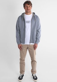 Sweat à capuche gris avec fermeture éclair et cordons, porté sur un T-shirt blanc avec un logo, associé à un pantalon beige et des baskets noires.