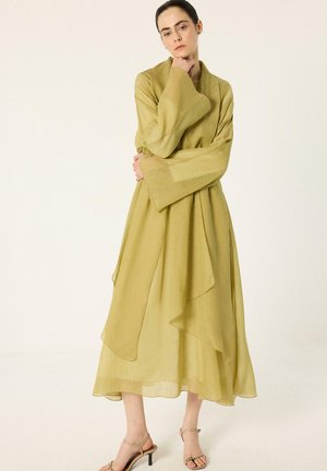 Femme portant une longue robe ample vert olive clair avec de larges manches et des sandales à talons hauts beige, se tenant devant un fond uni.