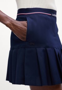 Námořnická plisovaná sukně s hladkou texturou. Obsahuje viditelnou kapsu a kontrastní červenobílý pruh na pásku. Přítomno logo Lacoste.
