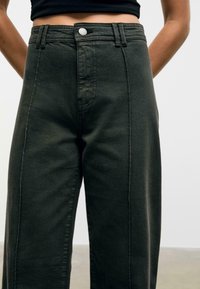 Pantaloni in denim verde scuro a vita alta con vestibilità dritta, cuciture a contrasto e chiusura con bottone frontale. Nessuna tasca visibile.