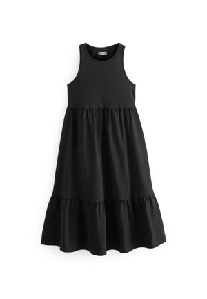 Personne portant une robe midi noire sans manches, une veste blanche boutonnée tombant des épaules, et des sandales noires à lanières, debout sur un sol gris.