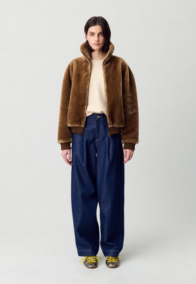 Chaqueta bomber de pelaje sintético marrón, suéter crema, jeans amplios azules, zapatillas gris y amarillo. Modelo de pie contra un fondo gris claro.