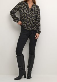 Frau in schwarzen, hochhackigen Knieboots, engen schwarzen Hosen und einem Langarmoberteil mit einem olivgrünen und schwarzen geometrischen Muster.