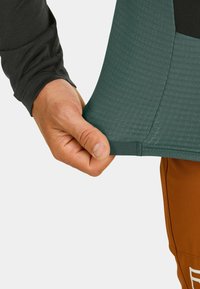 Langarmhemd aus strukturiertem dunkelgrauem Stoff mit grünem Saum. Eine Hand zieht an der Kante des Hemdes. Braune Hosen sind teilweise sichtbar.