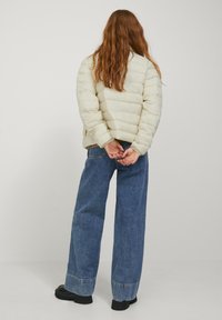 Pufferjack in lichtcrème met een gewatteerde textuur, gecombineerd met blauwe denim jeans met wijde pijpen en zwarte chunky laarzen. Achteraanzicht.