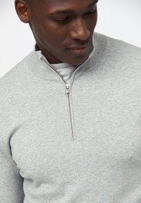 Hellgrauer Reißverschluss-Hoodie mit hohem Kragen, ausgestattet mit einem silbernen Reißverschluss und einem weichen, strukturierten Stoff. Schlichtes Design mit minimaler Detailierung.
