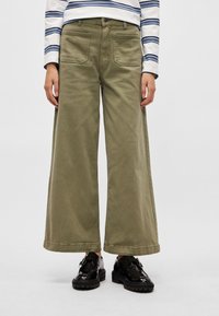 Ελιά πράσινο παντελόνι με φαρδιά γραμμή από βαμβάκι, με μπροστινές τσέπες και ακατέργαστο τελείωμα. Συνδυάζεται με λαμπερά μαύρα παπούτσια και ριγέ μακρυμάνικο πουκάμισο.