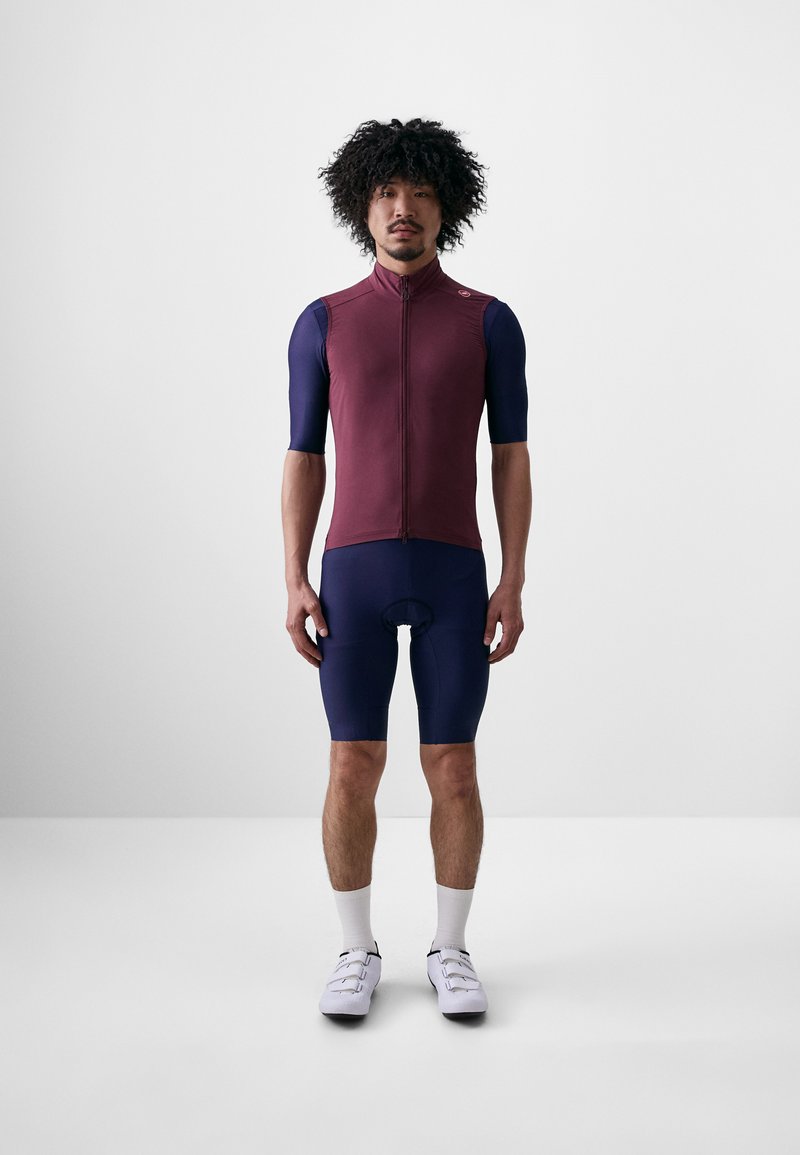 Maillot de cyclisme à manches courtes et zippé en bourgogne et bleu marine, associé à un short bleu marine. Le tissu semble lisse avec un ajustement près du corps. Chaussures de cyclisme blanches.
