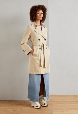 Cappotto trench doppiopetto beige con ampi rever e cintura in vita, abbinato a una gonna maxi in denim con spacco frontale e sneakers bianche.