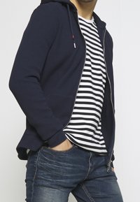 Námořnicky modrá mikina na zip s texturovanou látkou, s kapucí a bočními kapsami, vrstvená přes bílé a černé pruhované tričko, doplněná modrými džínami.
