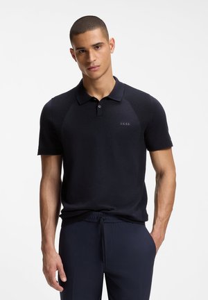 Νέος άνδρας με κοντά μαλλιά, που φοράει σκούρο μπλε πόλο μπλουζάκι και ταιριαστό παντελόνι, στέκεται με το ένα χέρι στην τσέπη μπροστά σε λευκό φόντο.