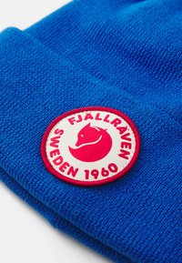 Mėlyna megzta kepurė su tekstūruotu paviršiumi. Yra apvalus baltas pleistras su raudonu lapės logotipu ir tekstu "FJÄLLRÄVEN SWEDEN 1960."