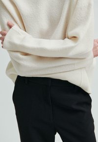 Osoba w kremowym, oversize'owym swetrze i czarnych spodniach, z założonymi za plecami rękami, na jednolitym jasnym tle.