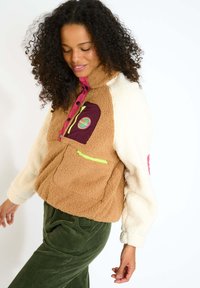 Giacca in pile marrone e crema con cerniera rosa e tasche. Presenta una toppa bordeaux e dettagli verde lime. Tessuto morbido, design corto.