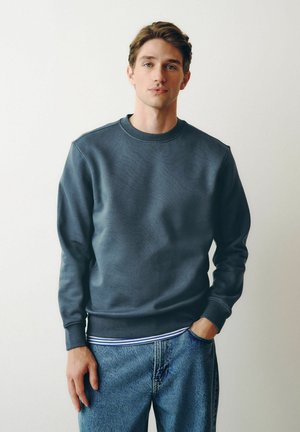 Young man wearing a dark gray sweatshirt and blue jeans, standing with one hand in pocket against a plain white background.
