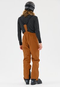 Bruine waterdichte sneeuwbroek met verstelbare banden, een relaxte pasvorm en zwarte accenten bij de enkels. Draagt een donker lange mouw top en een helm.