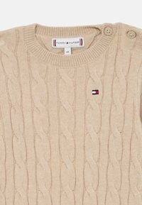 Pull en tricot beige en câble, fabriqué dans un tissu doux, avec un col rond et deux boutons sur l'épaule, accompagné d'un petit patch logo.