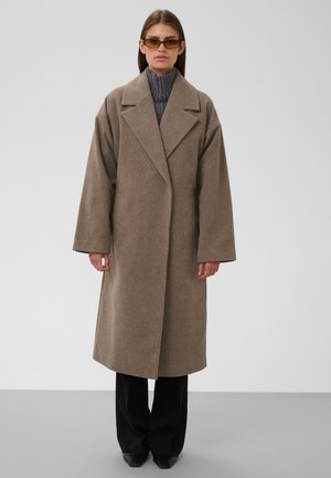 Vrouw met een oversized lichtbruine jas, grijze coltrui, zwarte broek met wijde pijpen, zwarte puntige schoenen en amberkleurige zonnebril, staand tegen een egale achtergrond.