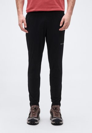 Man wearing black Columbia athletic pants and brown hiking shoes, standing with hands relaxed at sides on a plain white background.