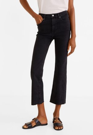 Persoon die zwart, hoog getailleerde cropped jeans met onafgewerkte zomen draagt, een wit T-shirt dat in de broek is gestopt, en zwarte slip-on sandalen met bandjes.