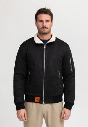 Man draagt een zwart jack met rits, witte kraag en beige broek, staand tegen een effen lichte achtergrond.