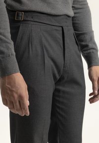 Hombre vistiendo pantalones plisados gris oscuro con detalle de cinturón y un suéter de manga larga gris oscuro a juego, con las manos relajadas a los lados.