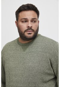 Crewneck genser i mørkegrønn strikket stoff med en teksturert halslinje. Designet har et subtilt melert mønster og ribbestrikkede mansjetter.