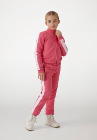 Pink tracksuit with white side stripes, made of soft material. Features a zip-up jacket and elastic waistband pants. White high-top sneakers.