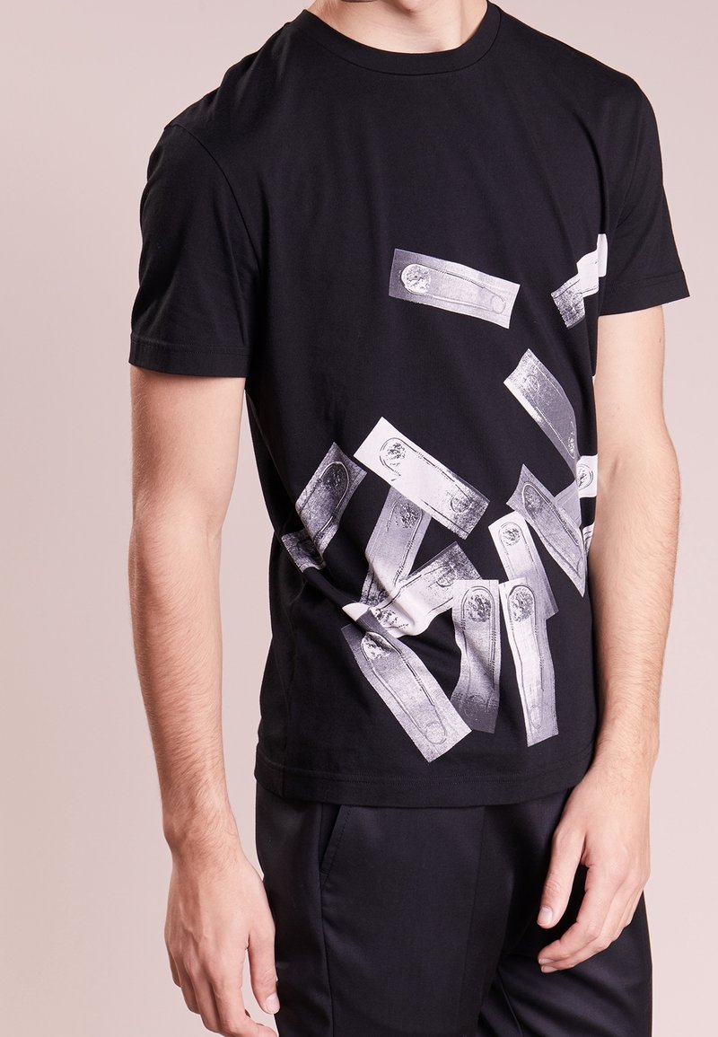 Homme portant un t-shirt noir avec plusieurs impressions graphiques rectangulaires blanches et grises dispersées sur le devant.