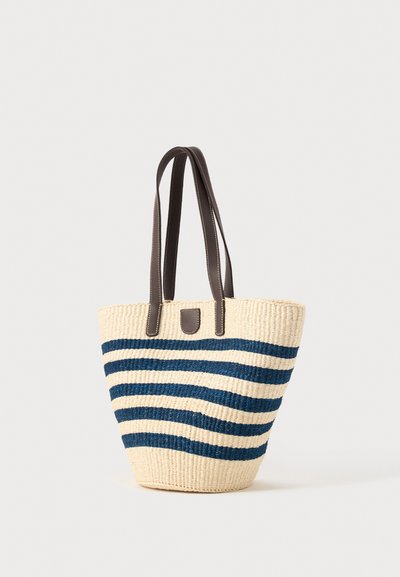 Πλεκτή μπεζ τσάντα tote με οριζόντιες σκούρες μπλε ρίγες και σκούρες καφέ δερμάτινες λαβές σε απλό ανοιχτό φόντο.