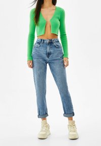 Vihreä ribattu lyhyt neuletakki syvällä V-aukolla, yhdistettynä korkea vyötäröiset sinisiin farkkuihin ja vaaleisiin lenkkareihin. Rentoa ja modernia tyyliä.