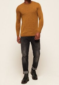 Pull à manches longues couleur moutarde avec un tricot texturé, associé à un jean foncé retroussé aux chevilles et des bottes en cuir noir.