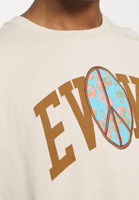 Béžové bavlněné tričko s velkým hnědým nápisem a symbolem míru zdobeným modrým květinovým vzorem. Kolačkový design.