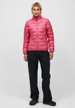 Lyshåret kvinde iført en pink quiltet jakke, sorte bukser og brune sko, stående foran en ensfarvet grå baggrund.