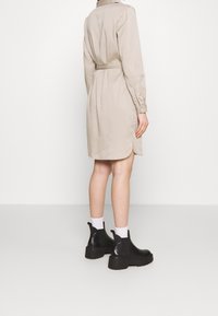 Beige mekko, jossa on pitkät hihat ja vyötäröllä vyö, pyöristetty helma. Yhdistetty mustiin nilkkureihin ja valkoisiin sukkiin.